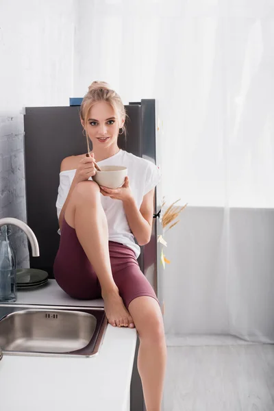 Young blonde woman sitting on kitchen table with bowl of breakfast — Stock Photo