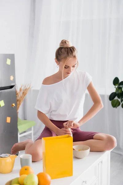 Donna bionda che apre una bottiglia di latte seduta sul tavolo della cucina vicino alla ciotola con cornflakes e frutta — Foto stock