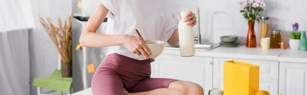 Cropped view of young woman holding bottle of milk and bowl in kitchen, panoramic concept — Stock Photo