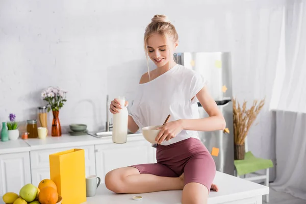 Sorrindo mulher loira sentada na mesa da cozinha e derramando leite em tigela — Fotografia de Stock