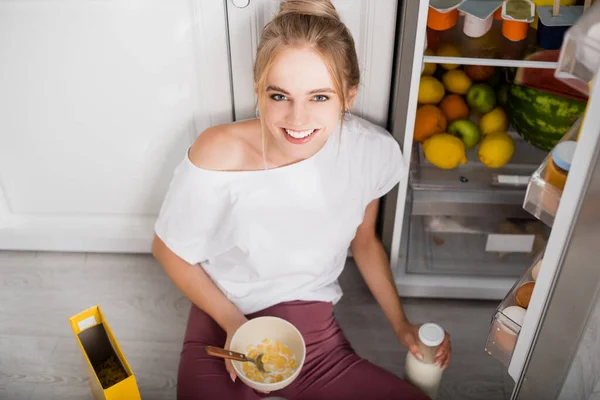 Blonde woman sitting on floor near opened fridge with bowl of cornflakes and bottle of milk — Stock Photo