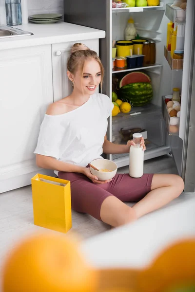 Selektiver Fokus einer jungen, fröhlichen Frau, die auf dem Boden neben dem geöffneten Kühlschrank mit einer Schüssel Cornflakes sitzt — Stockfoto