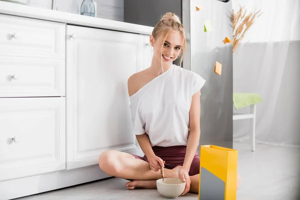 Sinnliche Frau lächelt in die Kamera, während sie mit Schüssel und Frühstück auf dem Boden sitzt — Stockfoto