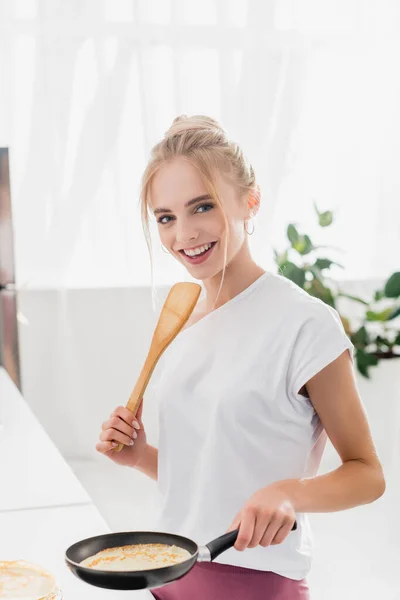 Mulher loira em t-shirt branca segurando espátula e frigideira com panqueca saborosa — Fotografia de Stock