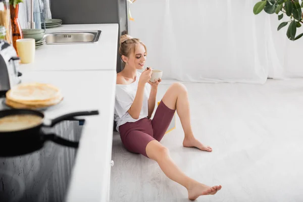 Selective focus of young woman in white t-shirt and shorts sitting on floor and eating breakfast from bowl — Stock Photo