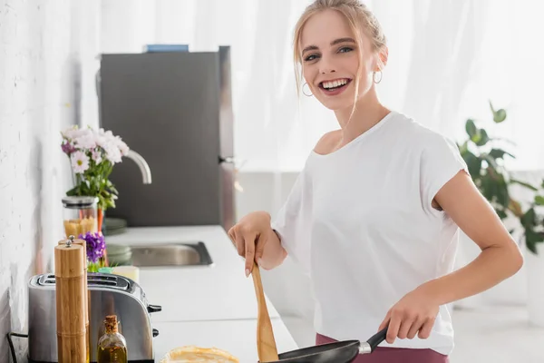 Excitada mujer rubia en camiseta blanca mirando a la cámara mientras cocina el desayuno - foto de stock