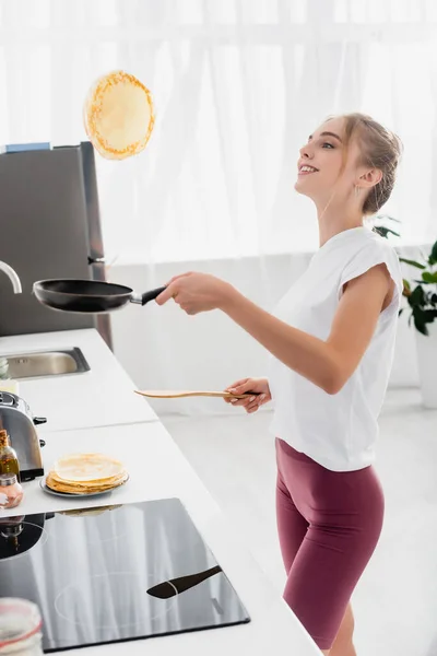 Young woman in white t-shirt and shorts throwing up pancake on frying pan while preparing breakfast — Stock Photo