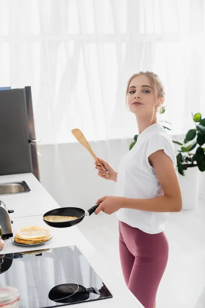 Giovane donna sensuale che tiene spatola e padella mentre prepara frittelle per la colazione — Foto stock