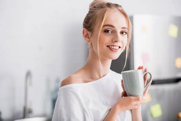 Freudige blonde Frau blickt in die Kamera, während sie mit einer Tasse Tee in der Küche steht — Stockfoto