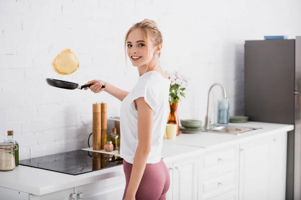 Молода блондинка дивиться на камеру, готуючи млинці на сковороді — стокове фото