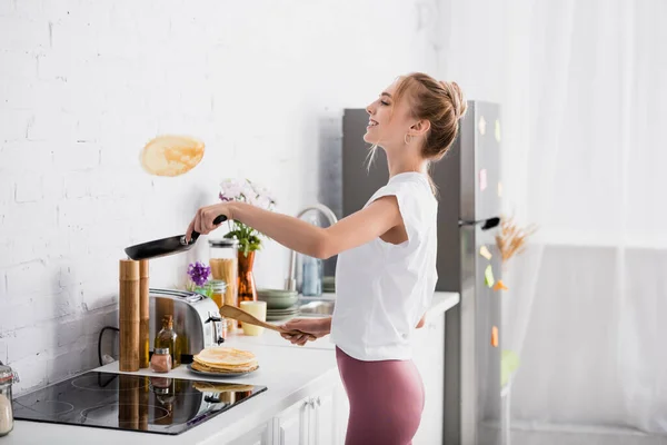 Side view of joyful blonde woman with frying pan and spatula preparing pancakes in kitchen — Stock Photo