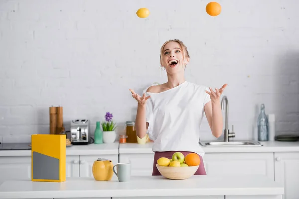 Animado jovem mulher malabarismo com frutas frescas enquanto está perto bule e xícara — Fotografia de Stock