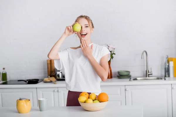 Excited blonde woman touching chin while covering eye with fresh apple in kitchen — Stock Photo