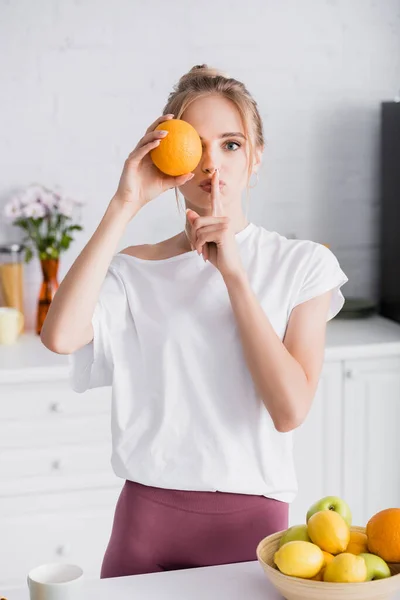 Mujer rubia joven oscureciendo el ojo con naranja fresca y mostrando gesto de silencio - foto de stock