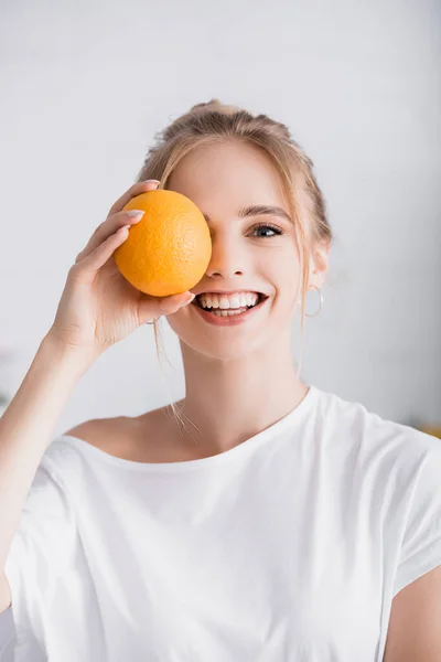 Happy blonde woman looking at camera while covering eye with whole ripe orange — Stock Photo