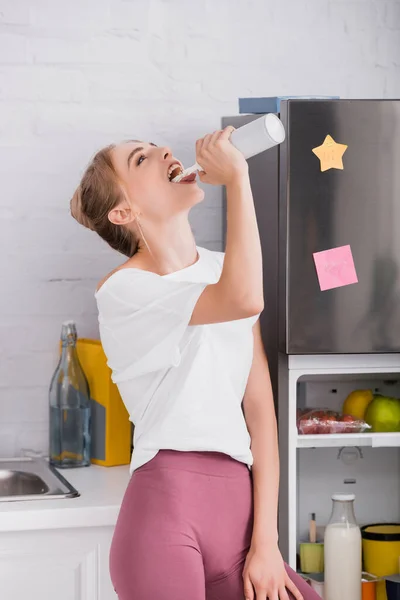 Young blonde woman eating whipped cream from bottle while standing near open fridge — Stock Photo