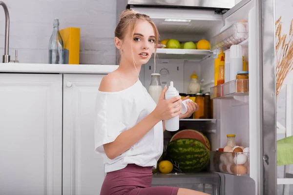 Mujer sensual en camiseta blanca sosteniendo botella de crema batida mientras está sentada cerca de la nevera abierta - foto de stock