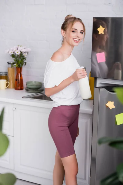 Glückliche junge Frau in weißem T-Shirt und kurzer Hose, die eine Flasche Milch in der Nähe des geöffneten Kühlschranks hält — Stockfoto