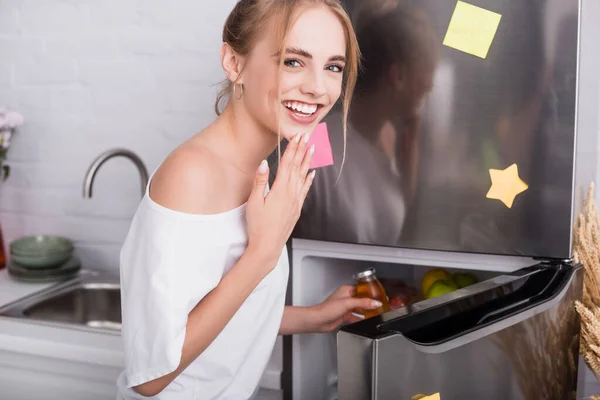 Allegra donna in t-shirt bianca che prende succo dal frigorifero mentre guarda la fotocamera — Foto stock