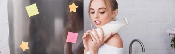Concepto horizontal de mujer rubia joven sosteniendo botella de leche cerca de la nevera - foto de stock