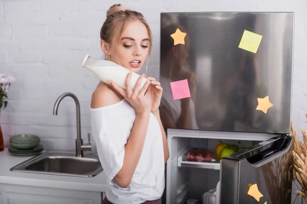 Молодая блондинка держит бутылку молока, стоя рядом с открытым холодильником — стоковое фото
