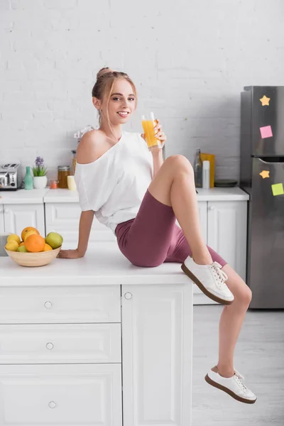 Mulher loira em camiseta branca e shorts sentados na mesa na cozinha com copo de suco de laranja — Fotografia de Stock
