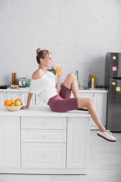 Vista lateral de mujer rubia en pantalones cortos y camiseta blanca sentada en la mesa de la cocina con vaso de jugo de naranja cerca de frutas frescas - foto de stock