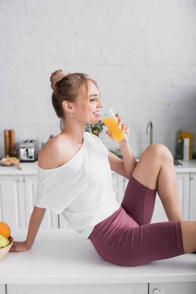 Side view of smiling woman in white t-shirt drinking orange juice while sitting on kitchen table — Stock Photo