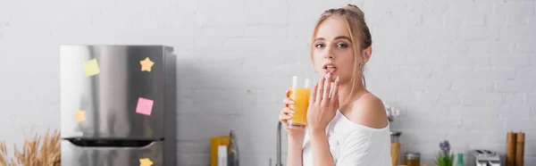 Horizontal concept of blonde woman holding glass of orange juice and touching lips in kitchen — Stock Photo