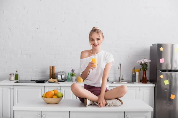 Joyful blonde woman holding orange juice while sitting on kitchen table with crossed legs — Stock Photo
