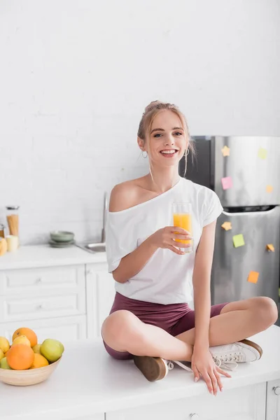 Mujer sonriente en camiseta blanca sentada en la mesa de la cocina con un vaso de jugo de naranja - foto de stock