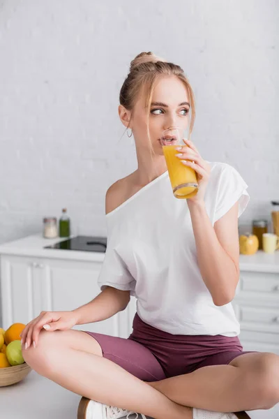 Blonde woman sitting on kitchen table with crossed legs and drinking orange juice — Stock Photo