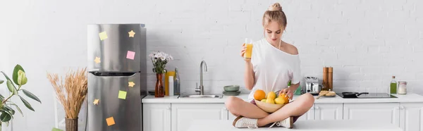 Concepto horizontal de mujer rubia joven sentada en la mesa de la cocina con frutas frescas y jugo de naranja - foto de stock