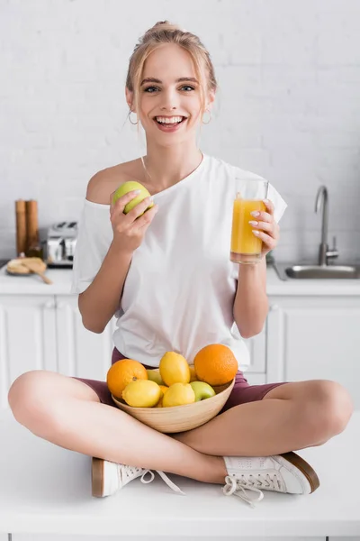 Pleased young woman smiling at camera while sitting on kitchen table with fruits and glass of orange juice — Stock Photo