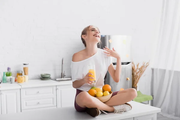 Mujer riendo en camiseta blanca sentada en la mesa de la cocina con frutas y un vaso de jugo de naranja - foto de stock