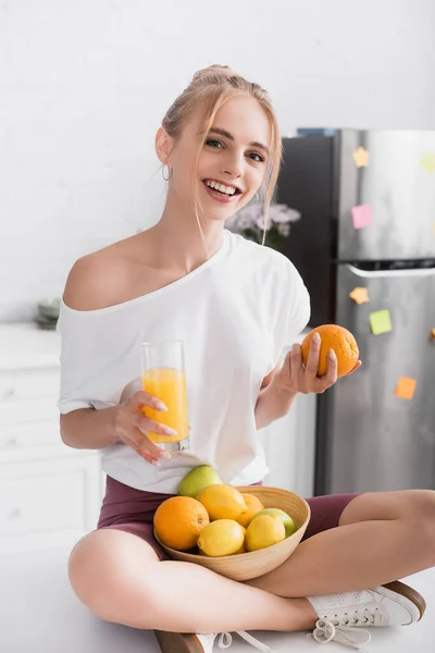 Jovem loira sentada na mesa da cozinha com suco de laranja e frutas frescas — Fotografia de Stock