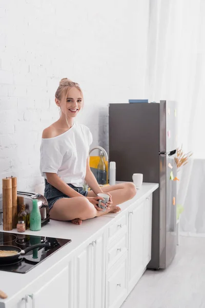 Радостная женщина сидит на кухонном столе со скрещенными ногами и смотрит в камеру — стоковое фото