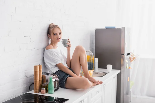 Sensual mujer joven en pantalones cortos sentado en la mesa de la cocina y mirando a la cámara - foto de stock