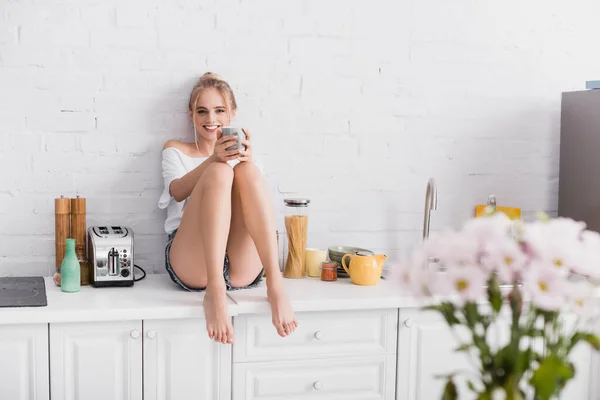 Jeune femme pieds nus en short assis sur la table de cuisine et regardant la caméra — Photo de stock