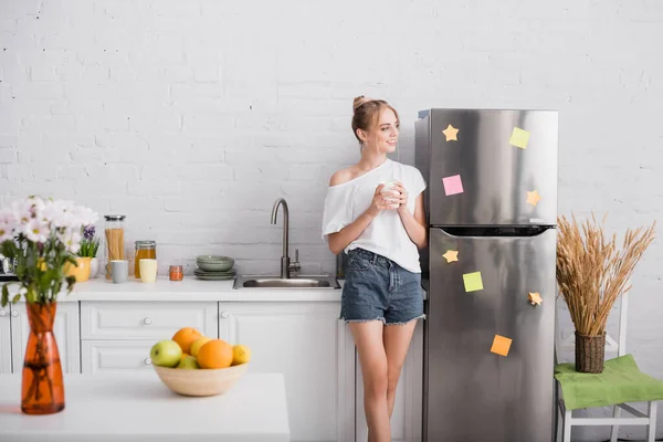 Foco seletivo da jovem mulher loira em t-shirt branca e shorts segurando copo enquanto está na cozinha — Fotografia de Stock