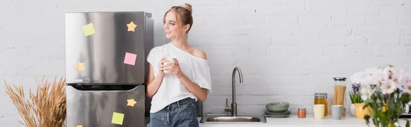 Панорамний знімок молодої жінки, що стоїть біля холодильника на кухні з чашкою чаю і дивиться в сторону — стокове фото