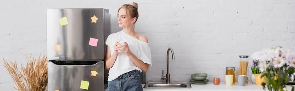 Plano panorámico de la joven mujer de pie cerca de la nevera en la cocina con taza de té y mirando hacia otro lado - foto de stock
