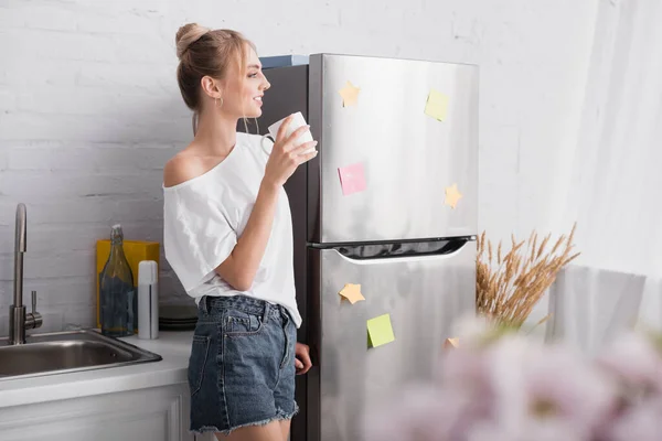 Selective focus of blonde woman in white t-shirt and denim shorts standing in kitchen with cup of tea — Stock Photo