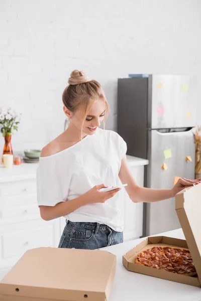 Jovem mulher em branco t-shirt mensagens no celular enquanto abre caixa com pizza na cozinha — Fotografia de Stock