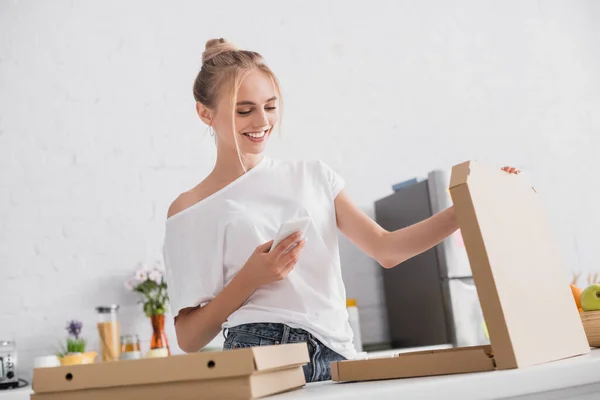 Pleased young woman messaging on smartphone while opening box with pizza — Stock Photo