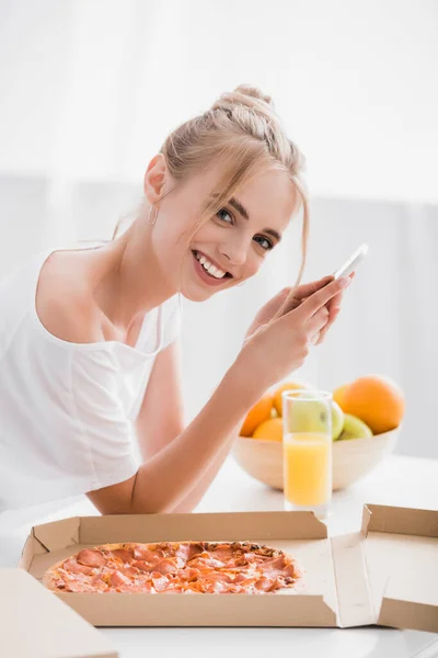 Mujer rubia complacida mirando a la cámara mientras chatea en el teléfono inteligente en la cocina - foto de stock