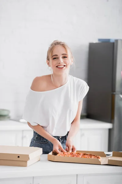 Mujer alegre mirando a la cámara mientras toma pizza de la caja en la cocina - foto de stock