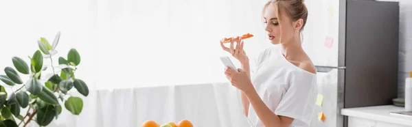 Website header of young woman chatting on smartphone while holding pizza in kitchen — Stock Photo