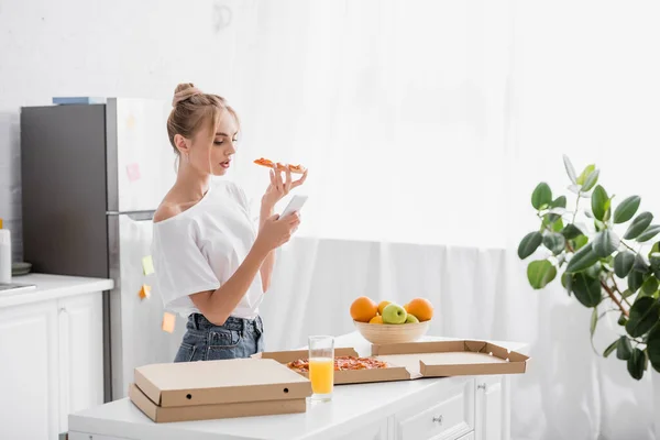 Mujer joven en camiseta blanca usando smartphone y sosteniendo pizza en la cocina - foto de stock