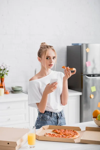 Blonde woman holding smartphone and pizza while looking at camera in kitchen — Stock Photo